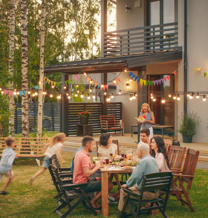 Family and friends enjoying an outdoor dinner in a backyard with string lights, colorful decorations and a wooden deck