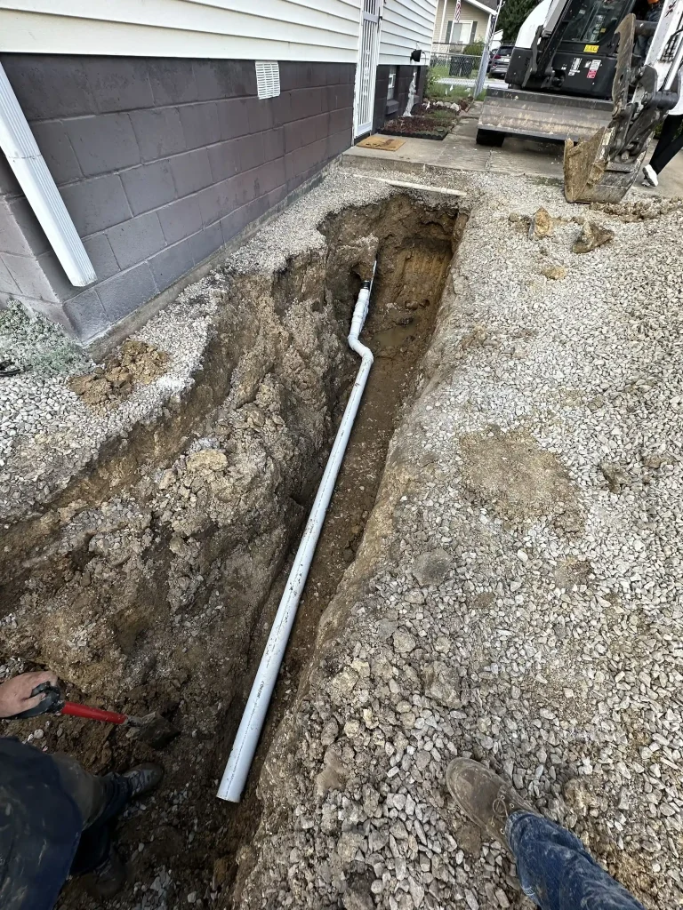 Excavated trench beside a residential foundation with white PVC drainage piping and construction equipment